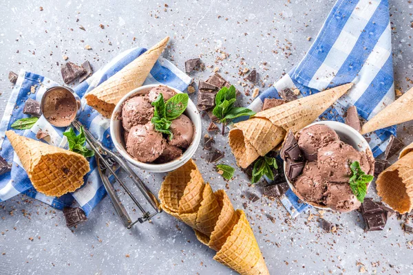 Homemade chocolate ice cream with chocolate pieces and shavings, and ice cream cones. In small white bowls on white grey stone table copy space