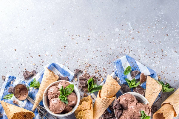 Homemade chocolate ice cream with chocolate pieces and shavings, and ice cream cones. In small white bowls on white grey stone table copy space