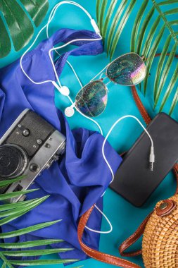 Summer vacation and travel concept. Beach accessories things - bag, hat, swimsuit, sunglasses, camera, tropical leaves on aquamarine, turquoise blue backdrop. Summer is coming trendy concept