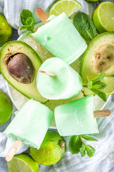 Homemade avocado popsicle. Vegan non-dairy ice cream. Avocado,lime and coconut healthy popsicle on grey stone background with fresh lime slices, avocados and ice