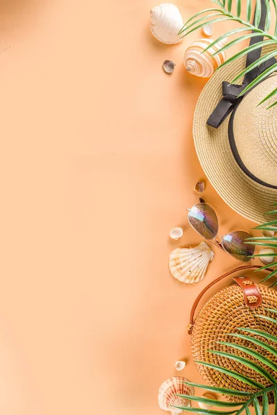 Trendy summer flat-lay with female fashion outfit - straw hat, bamboo bag, sunglasses, on peach background with  palm branches, top view, copy space, flat lay. Summer fashion, holiday concept