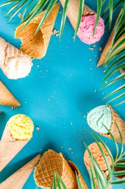 Dondurmalı waffle külahında çeşitli parlak renkli dondurma seti. Çikolatalı, yabanmersinli, şam fıstıklı, trend turkuaz arka planda ve tropikal palmiye yapraklı.