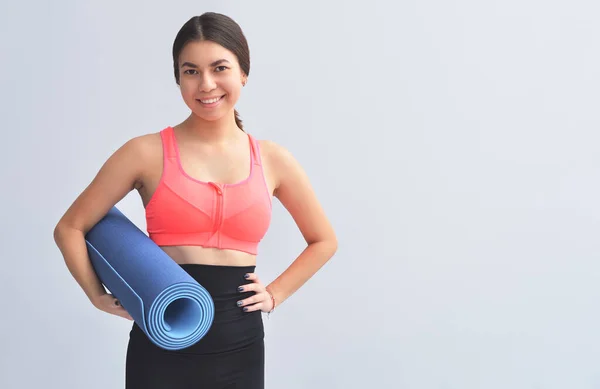 Elinde yoga minderi tutan ve gri arka planda egzersiz yapan güler yüzlü kız..