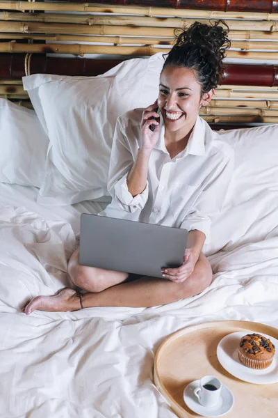Όμορφη γυναίκα με τηλέφωνο και φορητό υπολογιστή στο κρεβάτι — Φωτογραφία Αρχείου