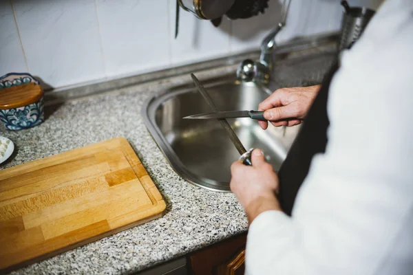 Hombre de 59 años afilar cuchillo en la cocina de su casa . — Foto de Stock