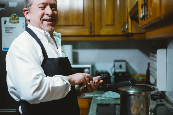 Homme de 59 ans avec smartphone dans la cuisine de sa maison . — Photo