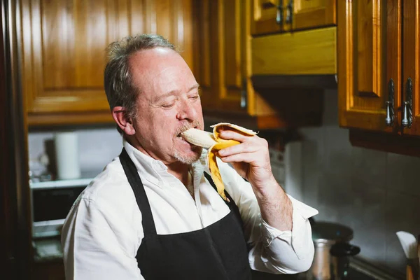 Hombre de 59 años come plátano en la cocina de su casa . — Foto de Stock