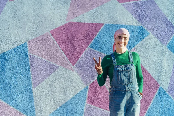 Woman with headscarf fights cancer on a colorful wall and showing victory shaped fingers