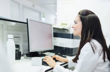 Hastane laboratuarındaki masada otururken tıbbi üniformalı bir kadının bilgisayar kullanışının yan görüntüsü.
