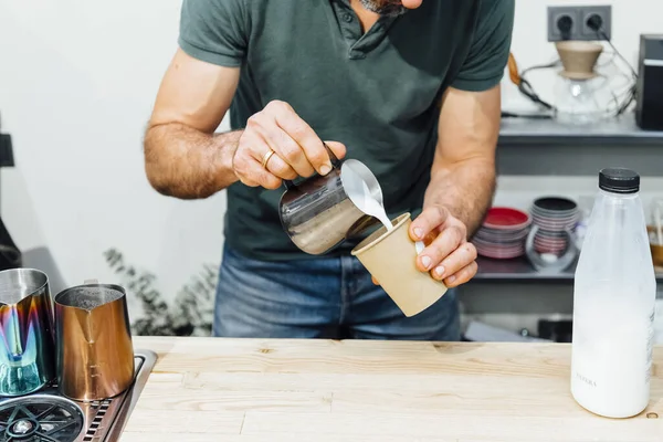 Close-up view of barista holding cup with espresso and pouring foamed milk over it while making cappuccino