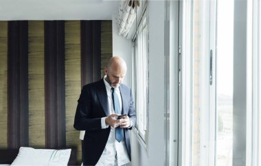 Pencerenin yanındaki akıllı telefonlu iş adamı.