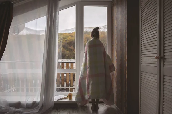 Girl stands near the window with mountain view in hotel room at morning time.