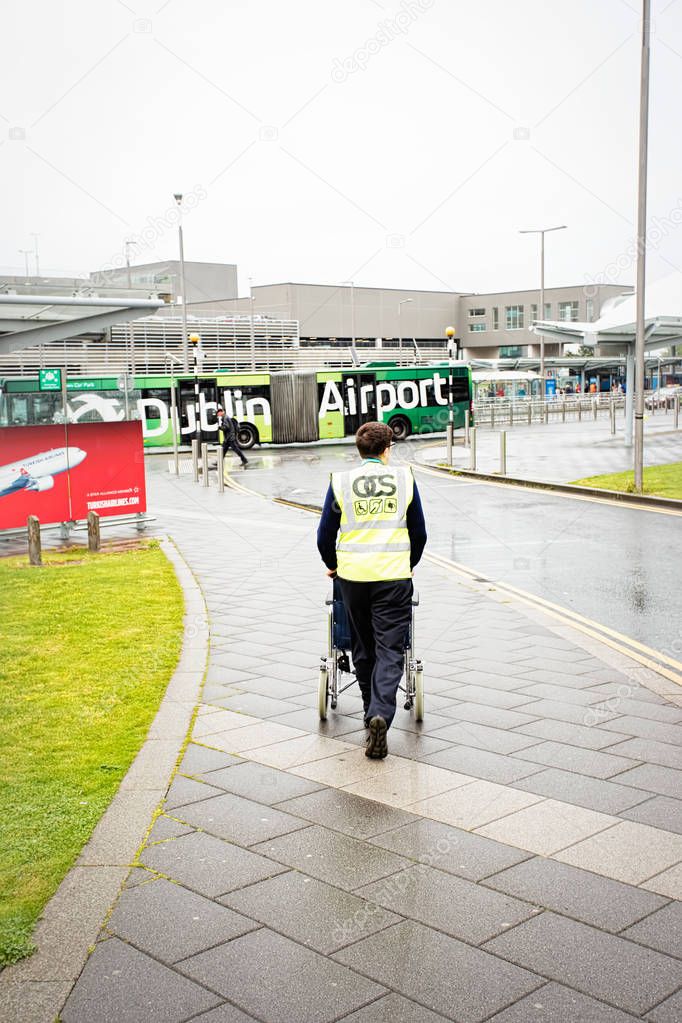 Dublin airport member staff pushing wheelchair for disabled passenger to airport building on summer rainy day, Dublin Airport, 14 August 2017