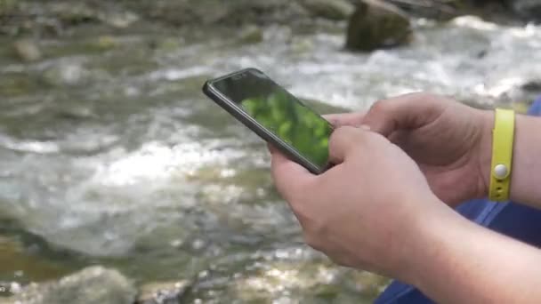 Surfen Sie im Internet oder tippen Sie Textnachrichten auf schwarzem Smartphone auf Bergbach-Hintergrund — Stockvideo