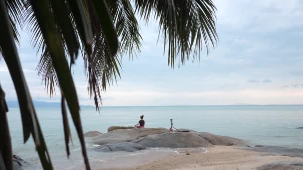 Macho se sienta en roca en frente de azul mar bajo azul cielo y hace vídeo — Vídeos de Stock