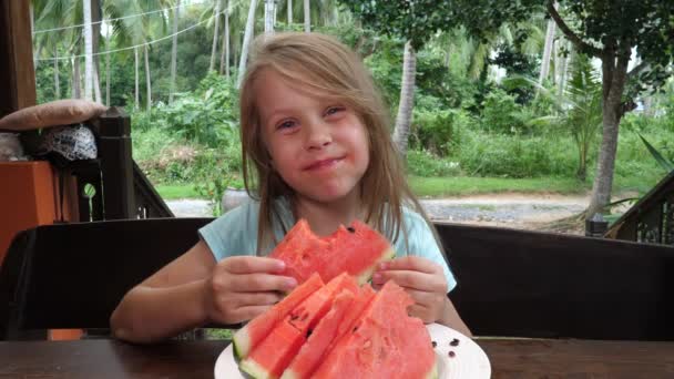 Petite fille souriante mignonne s'assoit à la table en bois à l'extérieur tenant et mordant tranche de melon d'eau rouge — Video