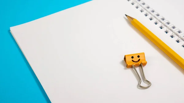 Bloc de notas de la escuela con lápiz amarillo y clip de carpeta de sonrisa naranja sobre fondo azul Imágenes de stock libres de derechos