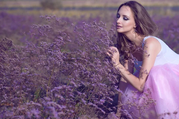 Young chic woman with artistic make up and long flying hair smelling violet flowers with closed eyes on the field.