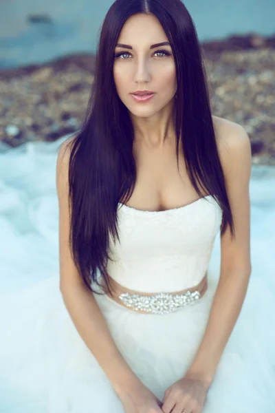 Retrato de la joven novia hermosa con el pelo largo negro en vestido de novia sentado a la orilla del mar y mirando con ternura recta . — Foto de Stock