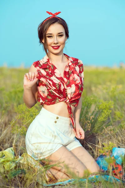 Hermosa joven con una sonrisa tentadora perfecta usando una blusa roja atada, pantalones cortos blancos y diadema sentados en el campo — Foto de Stock