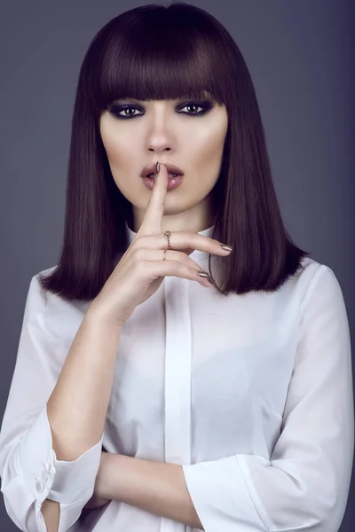 Portrait of gorgeous young dark-haired woman with provocative make up and expressive eyes looking straight and holding her forefinger close to the mouth with silence gesture — Stock Photo, Image
