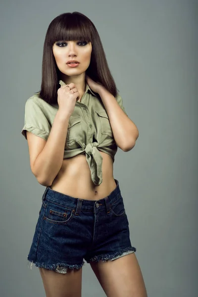 Portrait of young gorgeous dark-haired girl with provocative make up wearing high waisted dark blue jeans shorts and khaki tied up shirt with short sleeves. — Stock Photo, Image