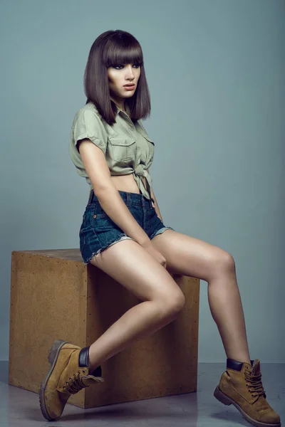Portrait of young gorgeous dark-haired model wearing high waisted dark blue jeans shorts, khaki shirt and brown boots sitting on the plywood cube and looking away — Stock Photo, Image