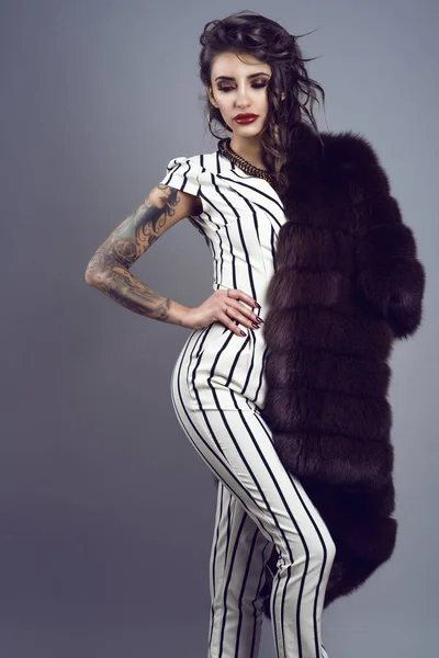 Portrait of young chic dark-haired tattooed lady wearing stylish striped overall with short sleeves and splendid necklace holding a furcoat on her shoulder — Stock Photo, Image