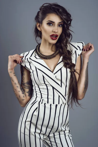 Portrait of young gorgeous dark-haired tattooed lady in striped overall and splendid necklace holding sleeves in her hands with surprised face expression — Stock Photo, Image