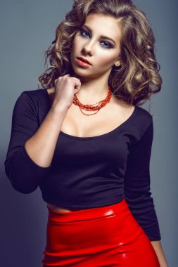 Portrait of young pretty model with long wavy hair wearing black top with three quarter sleeve and boat neckline and red leather skirt holding red beads on her neck clipart