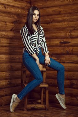 Portre genç güzel esmer model giyen sıska yüksek belli kot pantolon, çizgili gömlek, gerdanlık ve ahşap dışkı günlük kabinde oturan altın spor ayakkabı