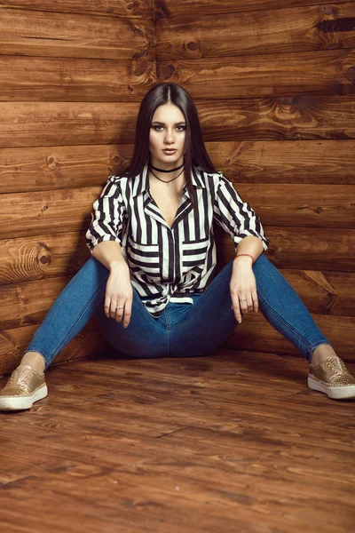 Retrato de una joven modelo sexy de cabello oscuro con pantalones vaqueros delgados de cintura alta, camisa atada a rayas con bolsillos, gargantilla y zapatillas doradas sentadas con las piernas abiertas en el suelo de madera en la cabaña de madera — Foto de Stock