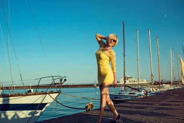 Retrato de linda loira de meia-idade sorrindo mulher usando amarelo apertado-cabido mini vestido de renda e óculos de sol dançando no cais do iate . — Fotografia de Stock