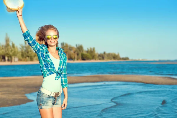 Retrato de una joven rubia delgada y sonriente con gafas de sol espejadas en forma de corazón, pantalones cortos de mezclilla y camisa azul con un sombrero en el brazo en un salto a la orilla del mar . — Foto de Stock