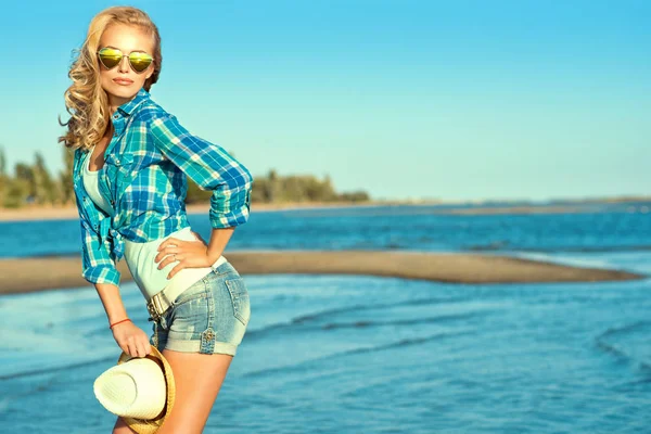 젊은 화려한 섹시 한 구릿빛 금발 미러 마음을 착용의 세로 모양의 선글라스, 데님 반바지와 그녀의 손에 파나마를 들고 해변에 서 있는 파란색 셔츠 체크. — 스톡 사진