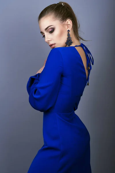 Portrait of young gorgeous model with ponytail and artistic make-up wearing electric blue open back dress and long delicate earrings keeping her hands at her breast — Stock Photo, Image