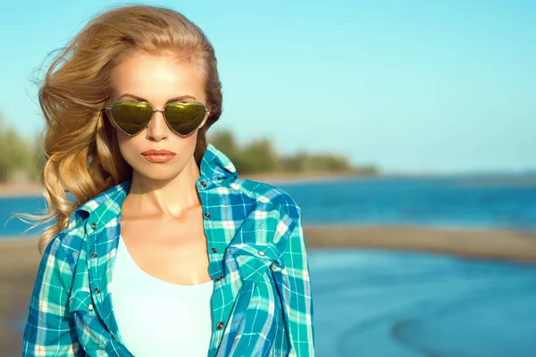 Retrato de la joven hermosa modelo rubia bronceada sexy con gafas de sol en forma de corazón espejado y camisa azul comprobado de pie en la playa, su cabello soplado por el viento —  Fotos de Stock