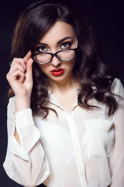 Όμορφη νεαρή γυναίκα με σκούρα κυματιστά μαλλιά και κόκκινα χείλη φορώντας λευκή μεταξωτή μπλούζα που αναζητούν ευθεία πάνω από τα γυαλιά της — Φωτογραφία Αρχείου