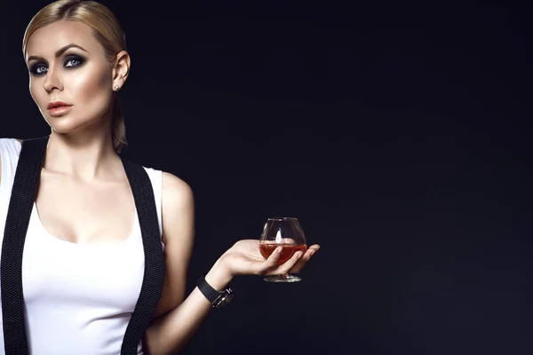 Portrait de modèle blond chic avec cheveux arrachés et maquillage des yeux fumé portant un haut blanc avec des bretelles sur les épaules et tenant un verre de brandy — Photo