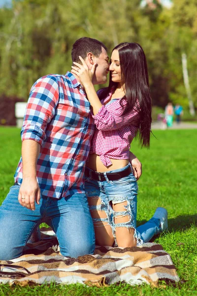Young beautiful happy loving couple in checked shirts and jeans standing on their knees on the green lawn and kissing