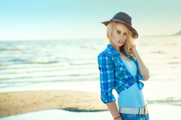 Портрет молодой роскошной сексуальной блондинки с дымчатыми глазами, макияж в черной войлочной шляпе и синей рубашке, стоящей на берегу моря, ее волосы развеваются ветром . — стоковое фото