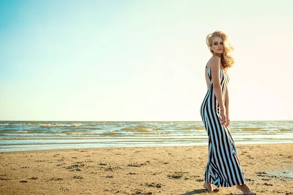 Retrato de uma mulher loira de cabelos compridos encantadora em vestido listrado preto e branco longo com costas nuas andando ao longo do litoral, seu cabelo ondulado soprado pelo vento — Fotografia de Stock