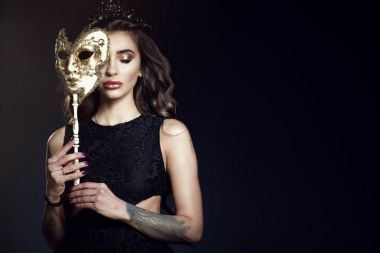 Beautiful tattooed lady with dark wavy shining hair and perfect make-up wearing lace dress and jewel black crown hiding the half of her face behind the Venetian mask and looking down clipart