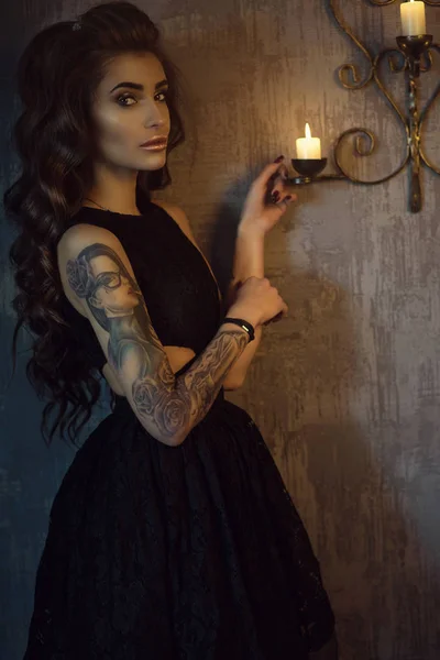 Portret chic młodych kobiety tatuażem z luksusowe dlugofalowe jedwabiste włosy i doskonały makijaż na sobie czarna Koronkowa sukienka z bokami Naga stojąc w ciemnym pokoju odrapane o świece — Zdjęcie stockowe