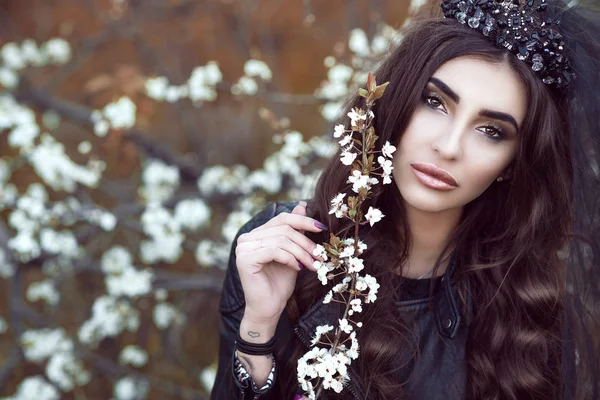 Close up portrait of a beautiful sad dark-haired young woman with perfect make up wearing black jewel crown with veil and leather jacket holding blossomed half faded branch — Stock Photo, Image