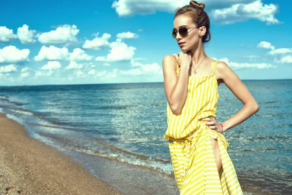 Retrato de una hermosa mujer delgada joven con el pelo updo usando vestido de verano holgado a rayas amarillas con abertura lateral y gafas de sol redondas espejadas de pie en la playa y mirando a un lado — Foto de Stock