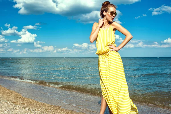 Retrato de uma bela jovem mulher sorridente com cabelo updo vestindo longo amarelo listrado vestido de verão folgado e redondo espelhado óculos de sol andando ao longo da beira-mar e olhando para o lado — Fotografia de Stock