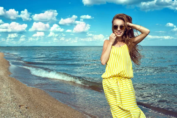 Retrato de uma bela jovem mulher sorridente com cabelos longos voando vestindo amarelo listrado vestido maxi verão folgado e redondo espelhado óculos de sol em pé à beira-mar — Fotografia de Stock