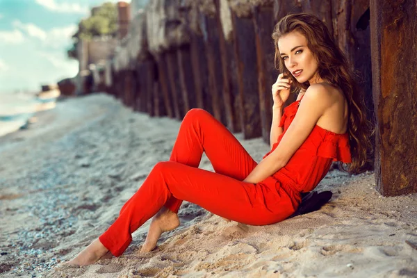 Retrato de uma jovem elegante encantadora com cabelos longos vestindo coral vermelho um macacão ombro sentado na praia nas velhas pilhas enferrujadas ao pôr do sol — Fotografia de Stock