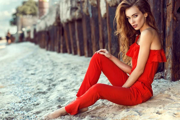 Retrato de una encantadora mujer joven elegante con el pelo largo con un mono rojo coral de un hombro sentado en la playa en las viejas pilas oxidadas al atardecer —  Fotos de Stock